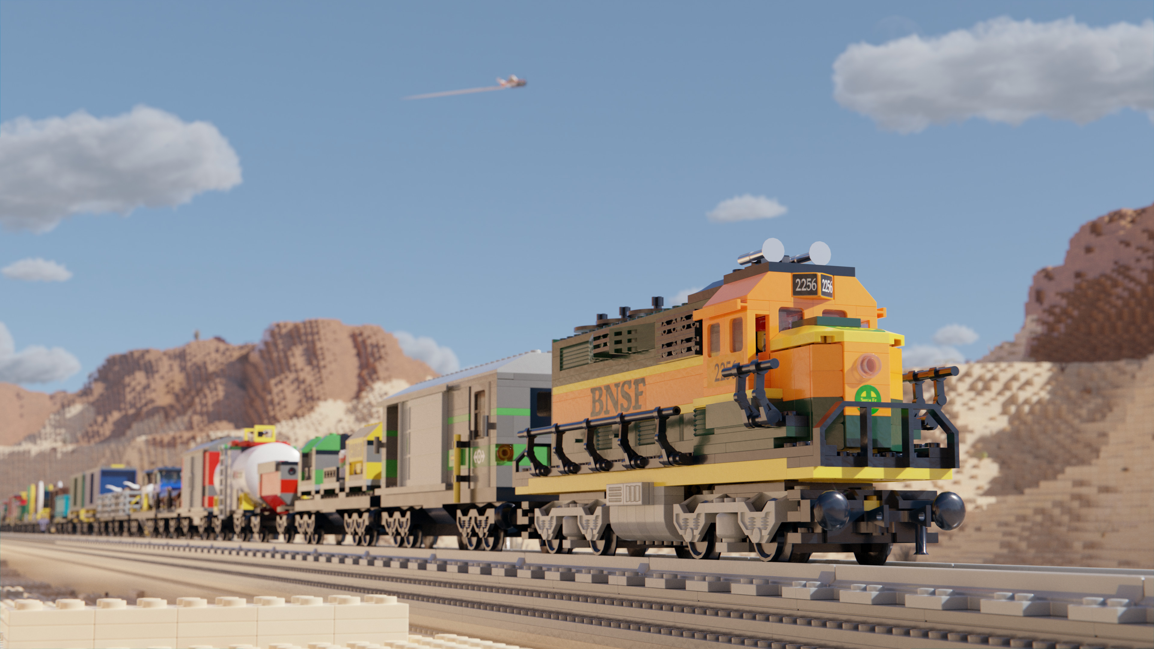 Image of a Lego train, travelling through a desert landscape on a sunny day, with some clouds in the sky and an airplane flying overhead.